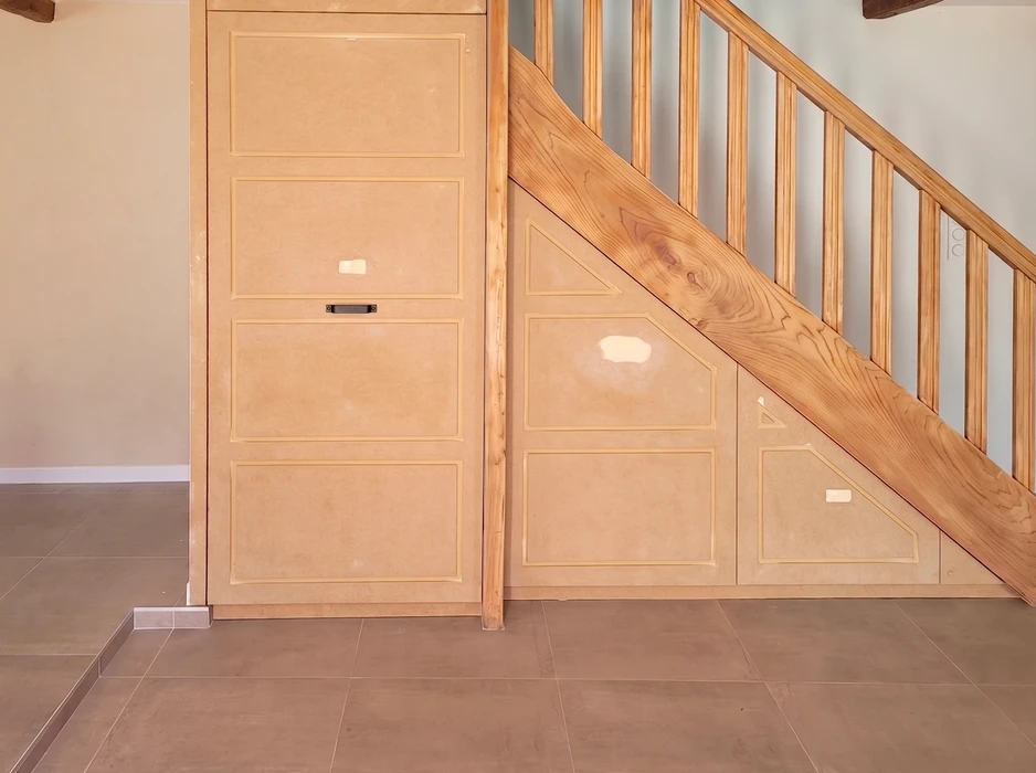Aménagement sous-escalier en MDF brut, comprenant plusieurs portes battantes à panneaux moulurés et tiroirs à poignée encastrée noire